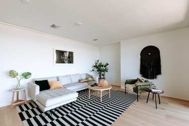 Living room with light wood floor, gray sofa, round coffee table, black-white rug, black textile wall hanging, and houseplants